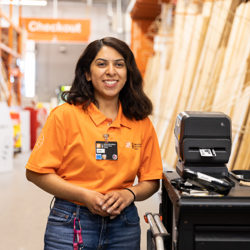 Young Latina woman in an orange polo shirt smiles as she leans against merchandising equipment.